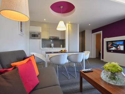 Nowoczesny pokój hotelowy z sofą, jadalnią, telewizorem i otwartą aneksem kuchennym w neutralnych i fioletowych tonacjach.