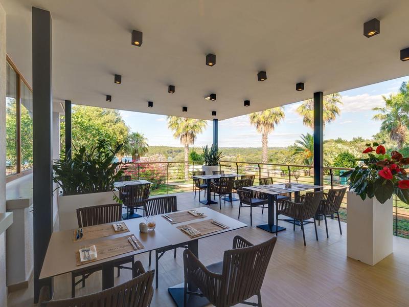 Zastřešená terasa s jídelními stoly a židlemi, obklopená zelenými rostlinami a výhledem na palmy.