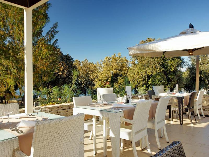 Slunečná venkovní terasa s bílými stoly a židlemi vedle zelených stromů a modré oblohy.