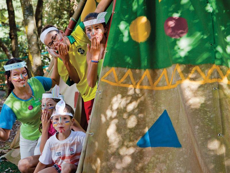 Děti s maskami zvířat hrají v lese vedle barevného stanu.