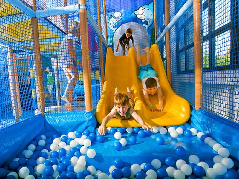 Děti sjíždějí ze žluté skluzavky do vnitřního herního prostoru plného modrých a bílých míčků.