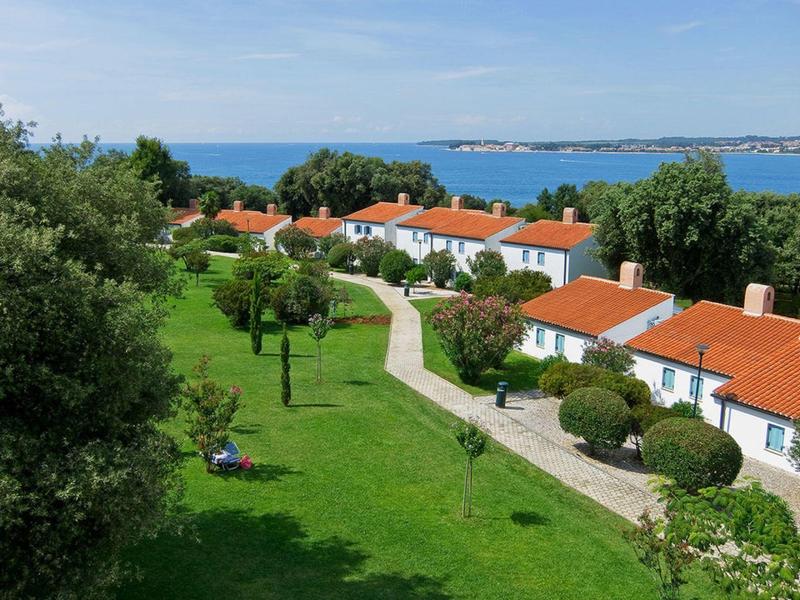 Rekreační domy s červenými střechami v zelené zahradě blízko moře pod modrou oblohou.