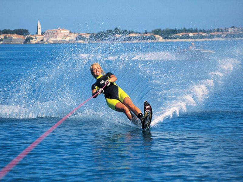 Osoba na vodních lyžích na modré vodě s pobřežním městem v pozadí pod jasnou oblohou.