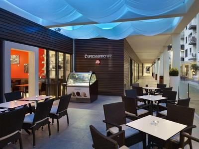 Moderní venkovní terasa restaurace se čtvercovými stoly a tmavými židlemi pod modře osvětlenou střechou.