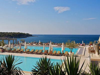 Pohled na hotelový bazén s lehátky a výhledem na moře za slunečného dne.