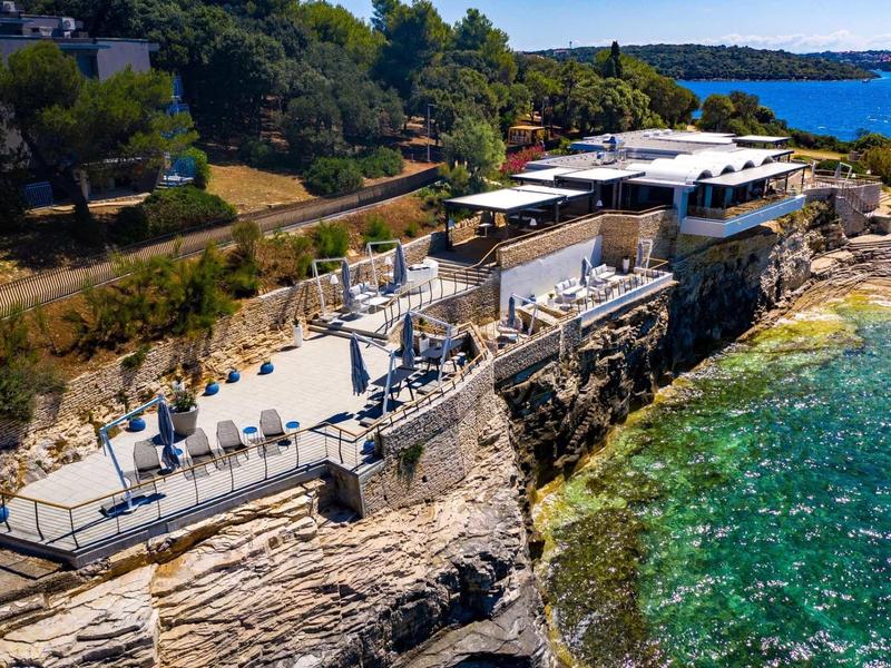 Küstenrestaurant auf Felsen mit Terrasse, Bäumen im Hintergrund und blau-grünem Meer bei Sonnenschein.