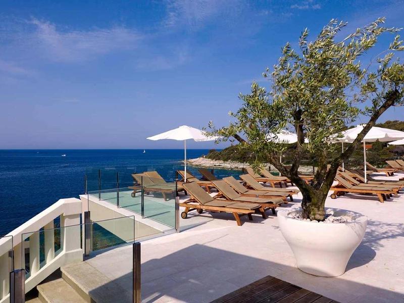 Terrasse mit Liegestühlen, Sonnenschirmen und einem großen Baum neben dem blauen Meer unter blauem Himmel.