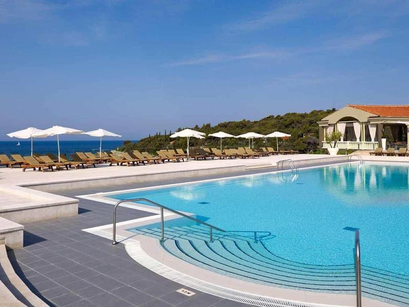 Großer blauer Pool mit Treppe, weißen Sonnenschirmen und rotdachigem Gebäude am Meer.