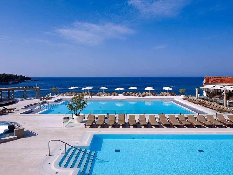 Großer Außenpool mit Liegestühlen, Sonnenschirmen und Blick auf das Meer unter blauem Himmel.