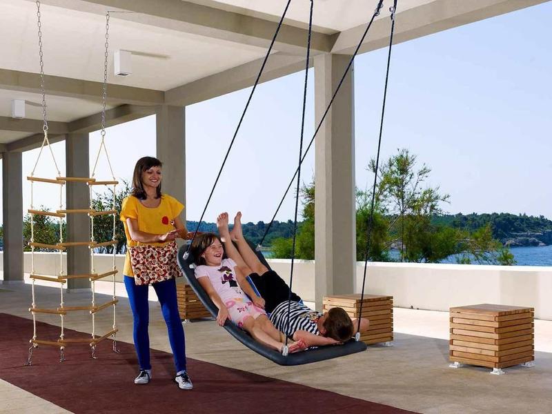 Ein Kind schaukelt auf einer Schaukel mit einer Frau, die daneben steht, unter einem modernen offenen Gebäude.