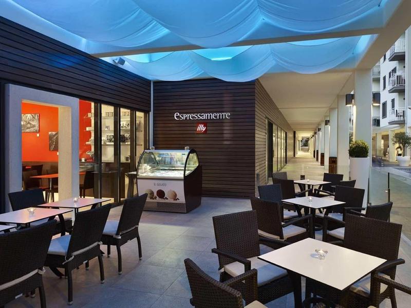 Moderner Außenbereich eines Cafés mit schwarzen Stühlen, weißen Tischen und blau beleuchtetem Dach.