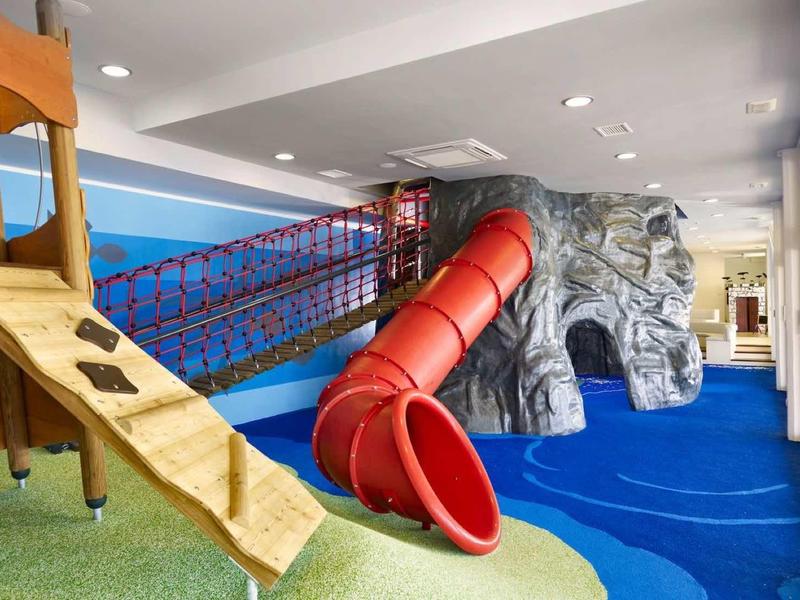 Innen-Spielplatz mit Rutsche, Klettergerüst und Kletterwand in bunten Farben und weichem Boden.