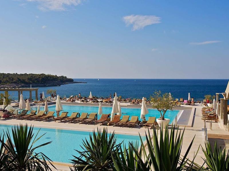 Blick auf zwei Pools mit weißen Liegestühlen, Palmen und dahinter blaues Meer unter blauem Himmel.