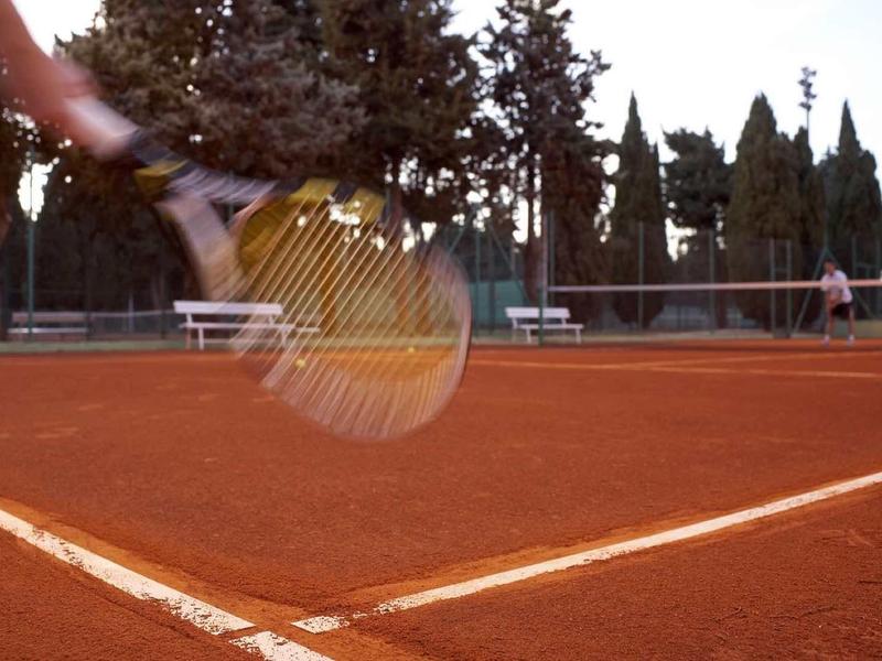 Tennisplatz mit rotem Sand und Spieler, der einen Ball mit gelbem Schläger schlägt.