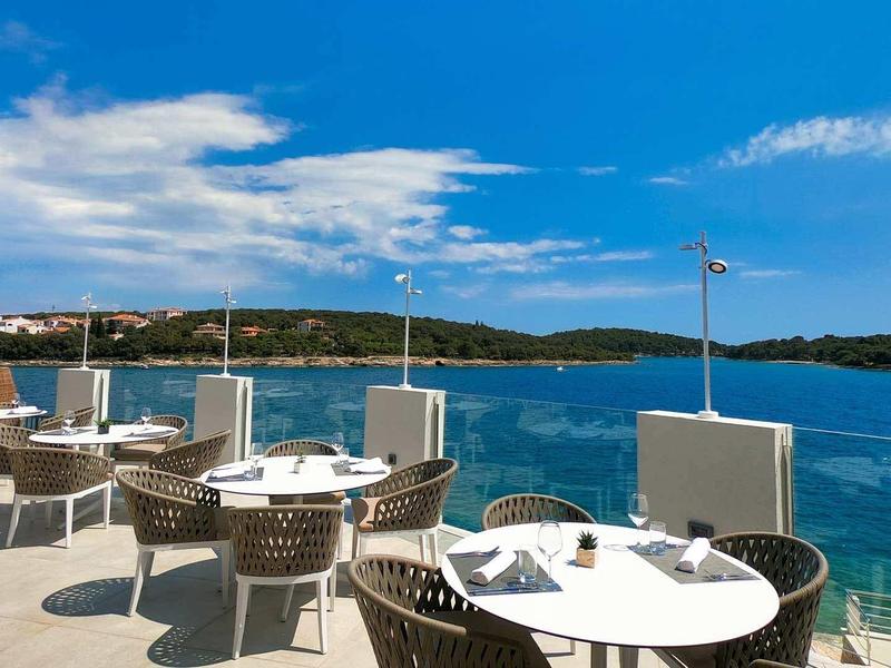 Terrasse mit weißen Tischen und Stühlen, Blick auf blaues Meer und grünen Küstenhorizont.