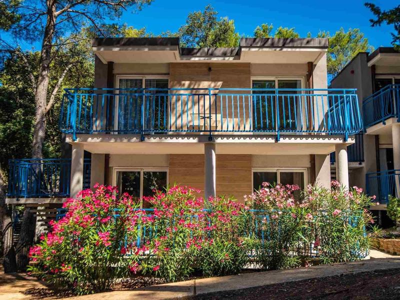 Zweistöckiges Haus mit blauer Balkonbrüstung, weißen Säulen und bunten Blumen vor dem Gebäude.