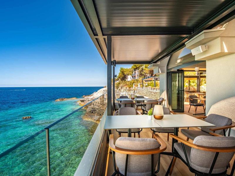 Balkon mit modernem Essbereich und Meerblick, türkisblaues Wasser und sonniger Himmel im Hintergrund.