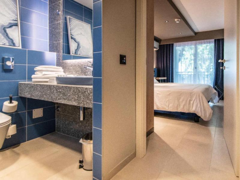 Moderner Hotelgang mit offenem Badezimmer mit blauen Fliesen und weißem Schlafzimmer mit Fenster.