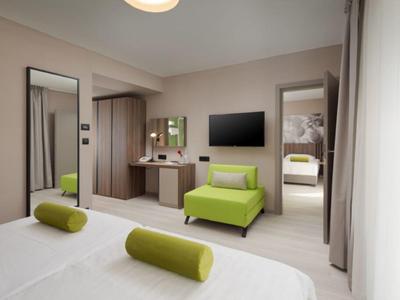 Moderně zařízený hotelový pokoj s manželskou postelí, zeleným křeslem, velkým zrcadlem a televizí.