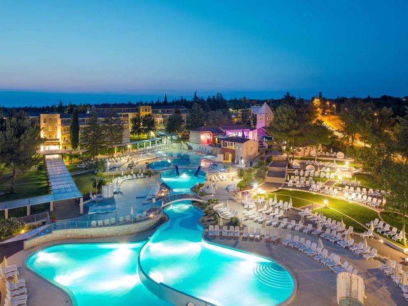 Osvětlené letovisko s několika bazény, lehátky a zahradními plochami za soumraku.