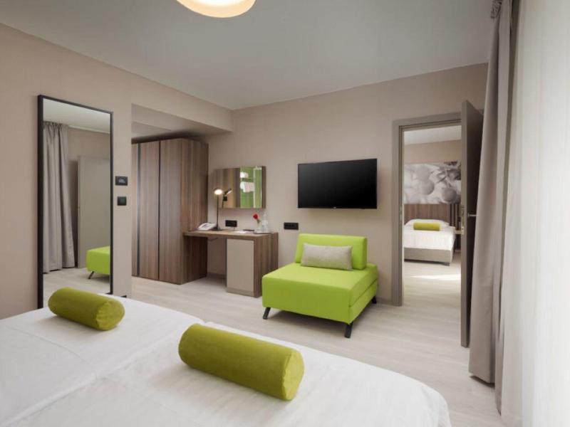 Moderně zařízený hotelový pokoj s manželskou postelí, zeleným křeslem, velkým zrcadlem a televizí.