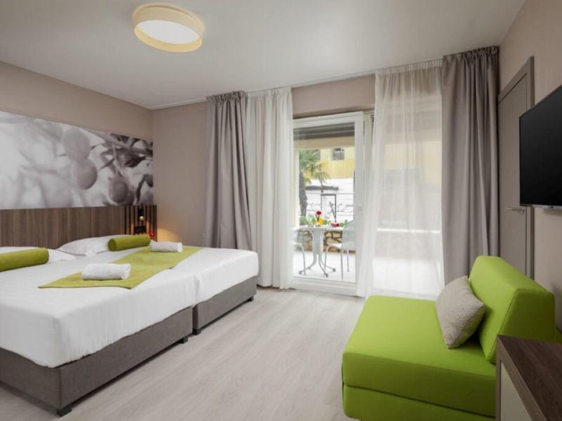Moderní hotelový pokoj se dvěma postelemi, zeleným křeslem a balkonem se stolem a židlemi.
