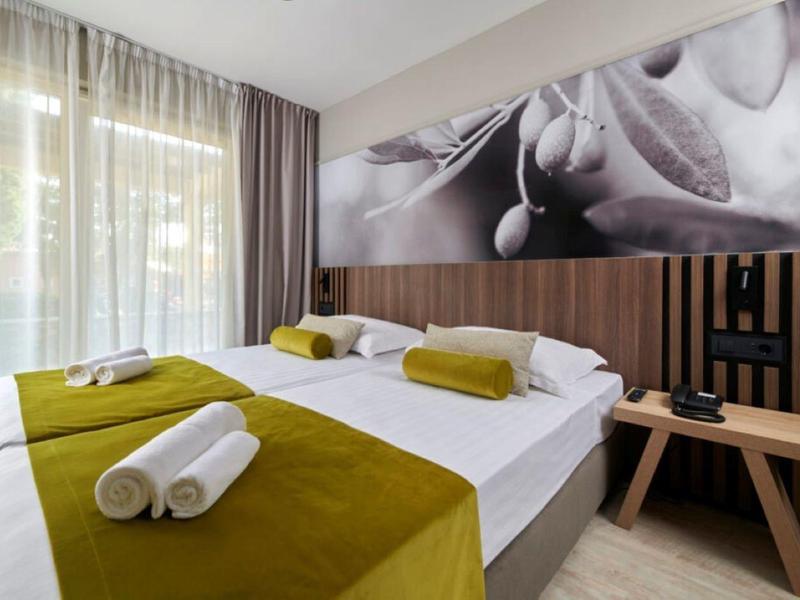 Moderní hotelový pokoj s manželskou postelí, zelenými dekami a velkými okny s závěsy.