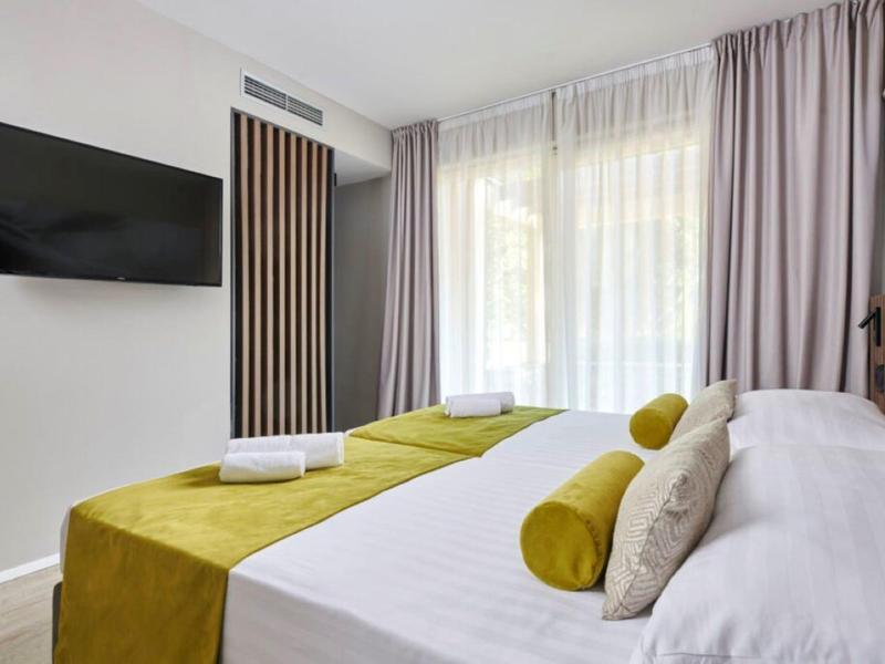 Moderní hotelový pokoj s manželskou postelí, žlutými akcenty a velkými okny s závěsy.