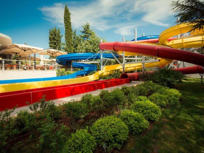 Barevné vodní skluzavky v venkovním bazénu se zelení a modrou oblohou.