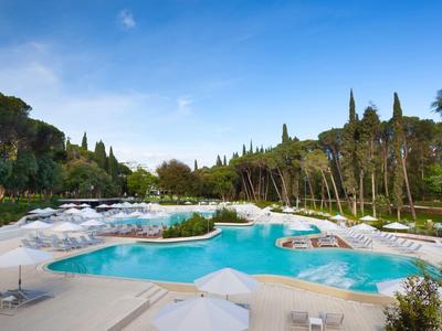 Großer Pool mit klarem Wasser, umgeben von Liegestühlen und grünen Bäumen unter blauem Himmel.