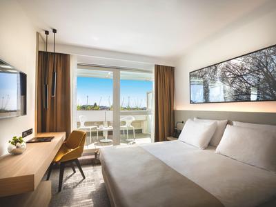 Schlafzimmer mit großem Bett, Schreibtisch, Stuhl, Wand-TV und Fenster mit Blick auf Hafen und Himmel.