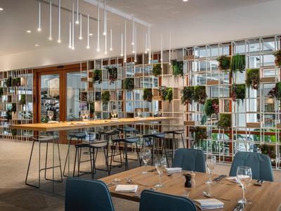 Moderner Gastraum mit Holzregalen, grünen Pflanzen und großen Fensterfronten in hellem Ambiente.