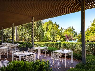 Terrasse mit weißen Stühlen und Tischen, umgeben von Grün und Bäumen, unter einem Holzdach.
