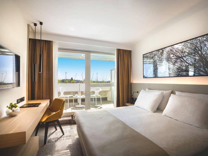 Schlafzimmer mit großem Bett, Schreibtisch, Stuhl, Wand-TV und Fenster mit Blick auf Hafen und Himmel.