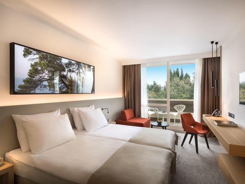 Helles Hotelzimmer mit zwei Betten, modernem Mobiliar und großem Fenster mit Blick auf Garten.