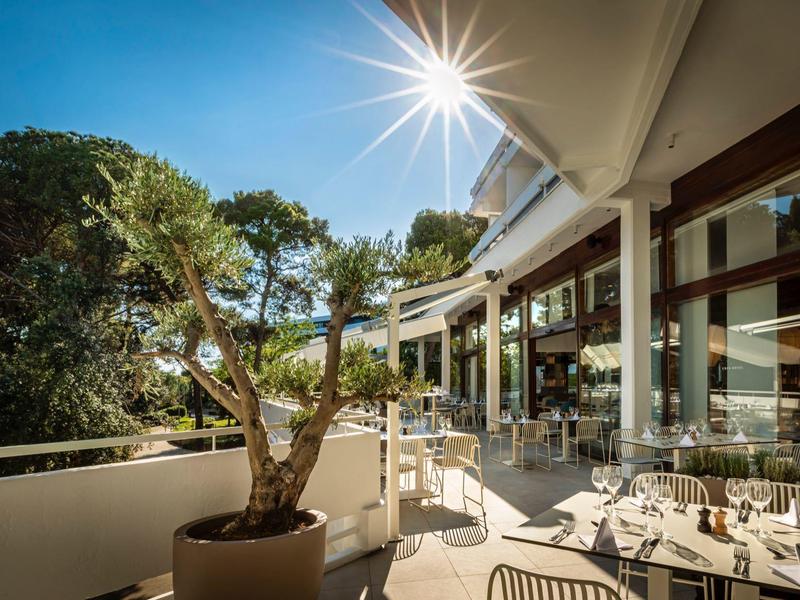 Sonniger Balkon mit Bäumen, weißen Stühlen und großer Glasfassade eines modernen Hauses.
