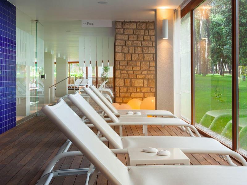 Moderner Ruheraum mit weißen Liegestühlen, Glaswand und Blick auf grüne Wiese und Bäume.