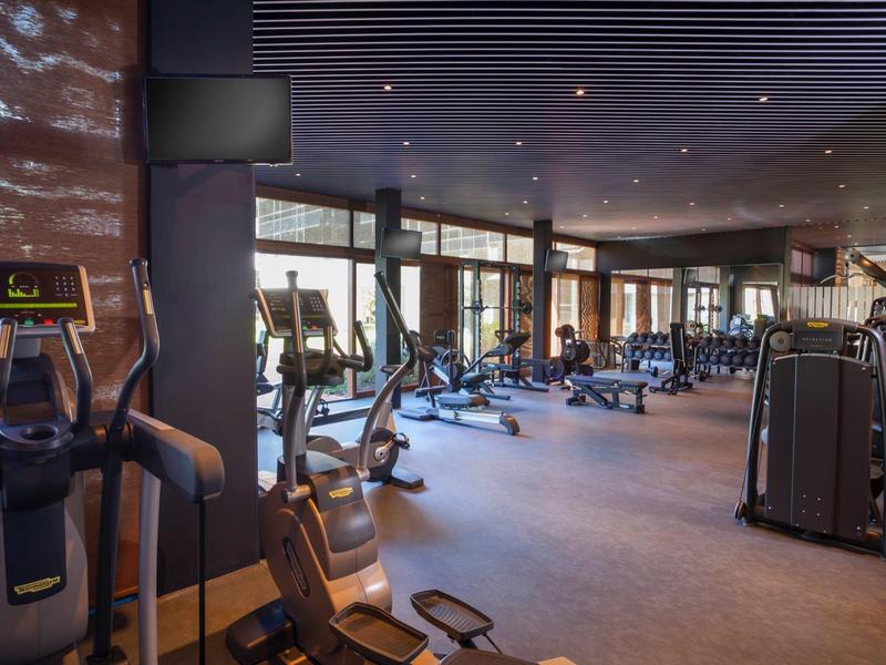 Großer, heller Fitnessraum mit modernen Geräten, Holzboden und großen Fenstern.