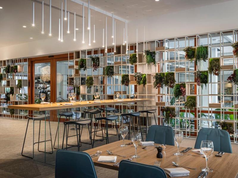 Moderner Gastraum mit Holzregalen, grünen Pflanzen und großen Fensterfronten in hellem Ambiente.