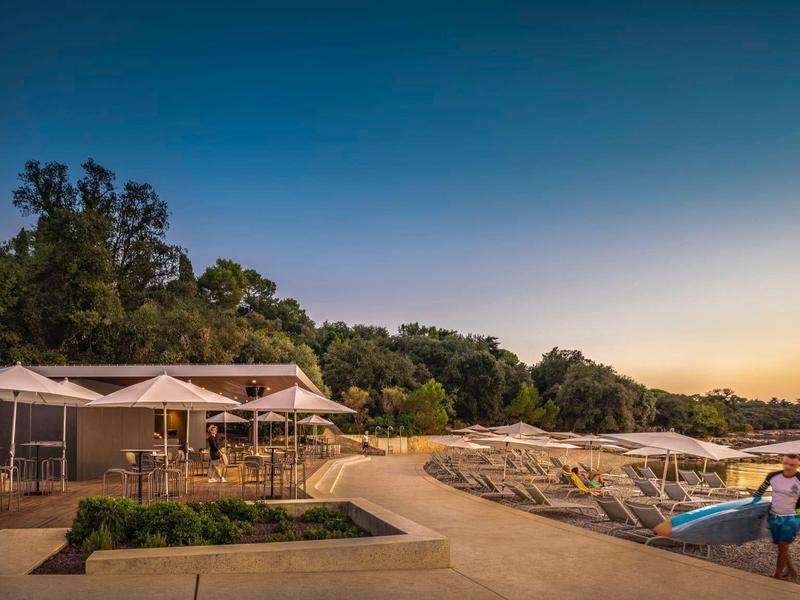 Sonniges Strandresort mit Liegestühlen, großen Sonnenschirmen und Bäumen im Hintergrund.