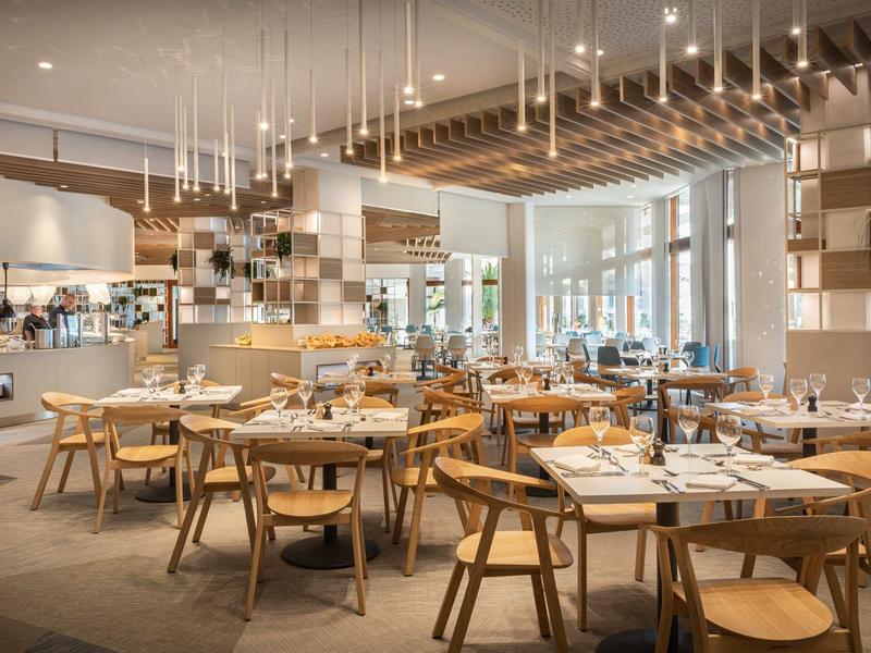 Modernes Restaurant mit Holzstühlen, runden Tischen, großen Fenstern und vielen Hängelampen.