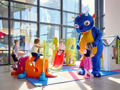 Kinder spielen in einem hellen, modernen Innenbereich mit einer Person im blauen Drachenkostüm.