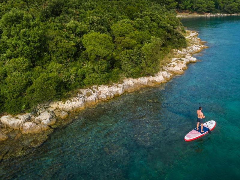 Eine Person steht auf einem Paddleboard auf klarem blauem Wasser neben einem bewaldeten, felsigen Ufer.