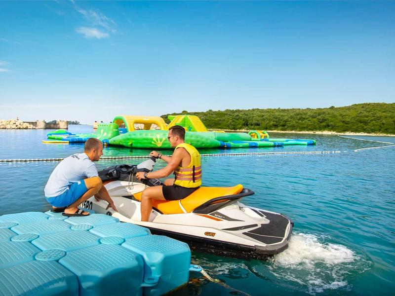 Zwei Männer sitzen auf einem Jetski auf blauem Wasser, im Hintergrund eine grüne Wasserrutsche.
