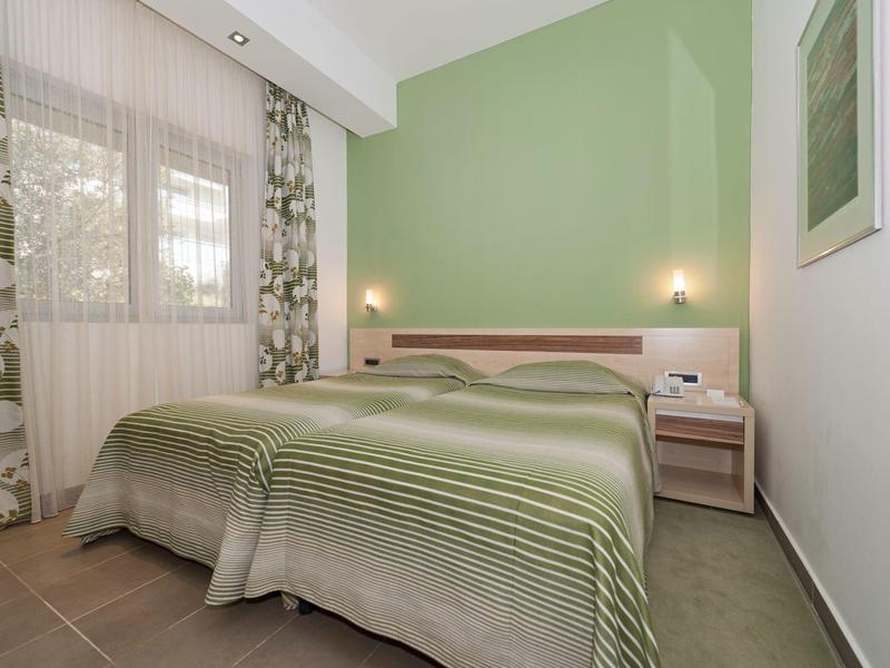 Hellgrünes Schlafzimmer mit Doppelbett, gestreifter Bettdecke, zwei Nachttischen und floralem Vorhang.