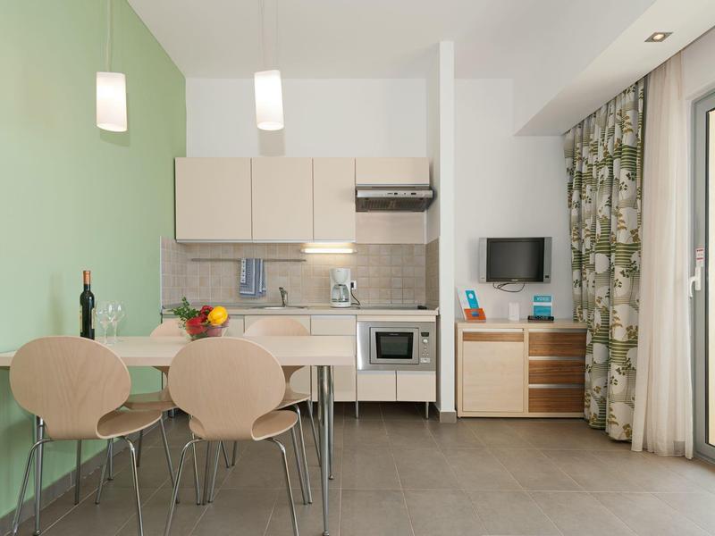 Kleine helle Küche mit grün gestrichener Wand, Esstisch, Stühlen und Vorhang am Fenster.