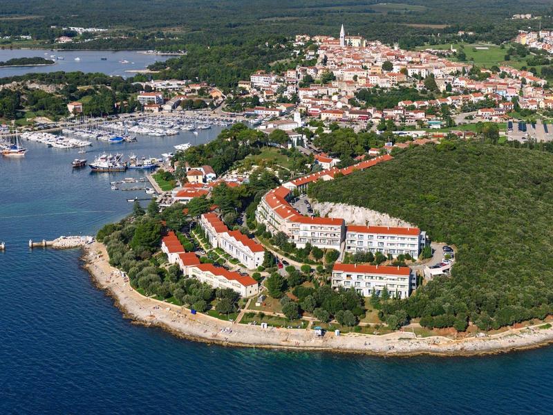 Luftaufnahme einer Küstenstadt mit Hafen, roten Dächern, grünen Wäldern und blauem Wasser.