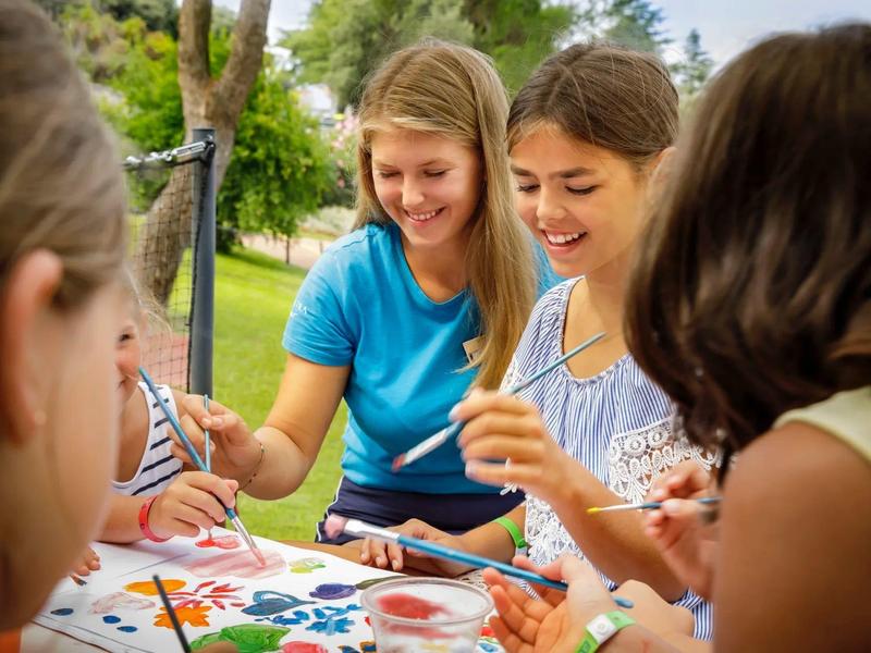 Kinder malen mit Pinseln bunte Bilder im Freien, umgeben von Grün und Sommerlicht.