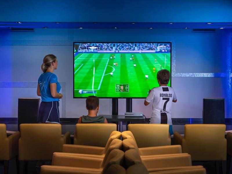 Zwei Kinder und ein Erwachsener schauen auf einem Sofa ein Fußballspiel auf großem Bildschirm.