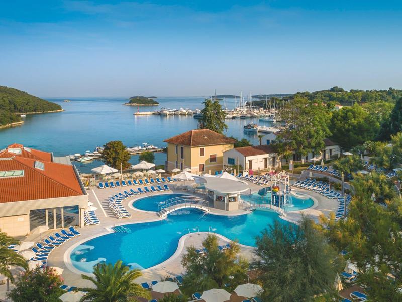 Hotelanlage mit geschwungenem Pool, vielen Liegen, Palmen und Blick auf das blaue Meer und Inseln.